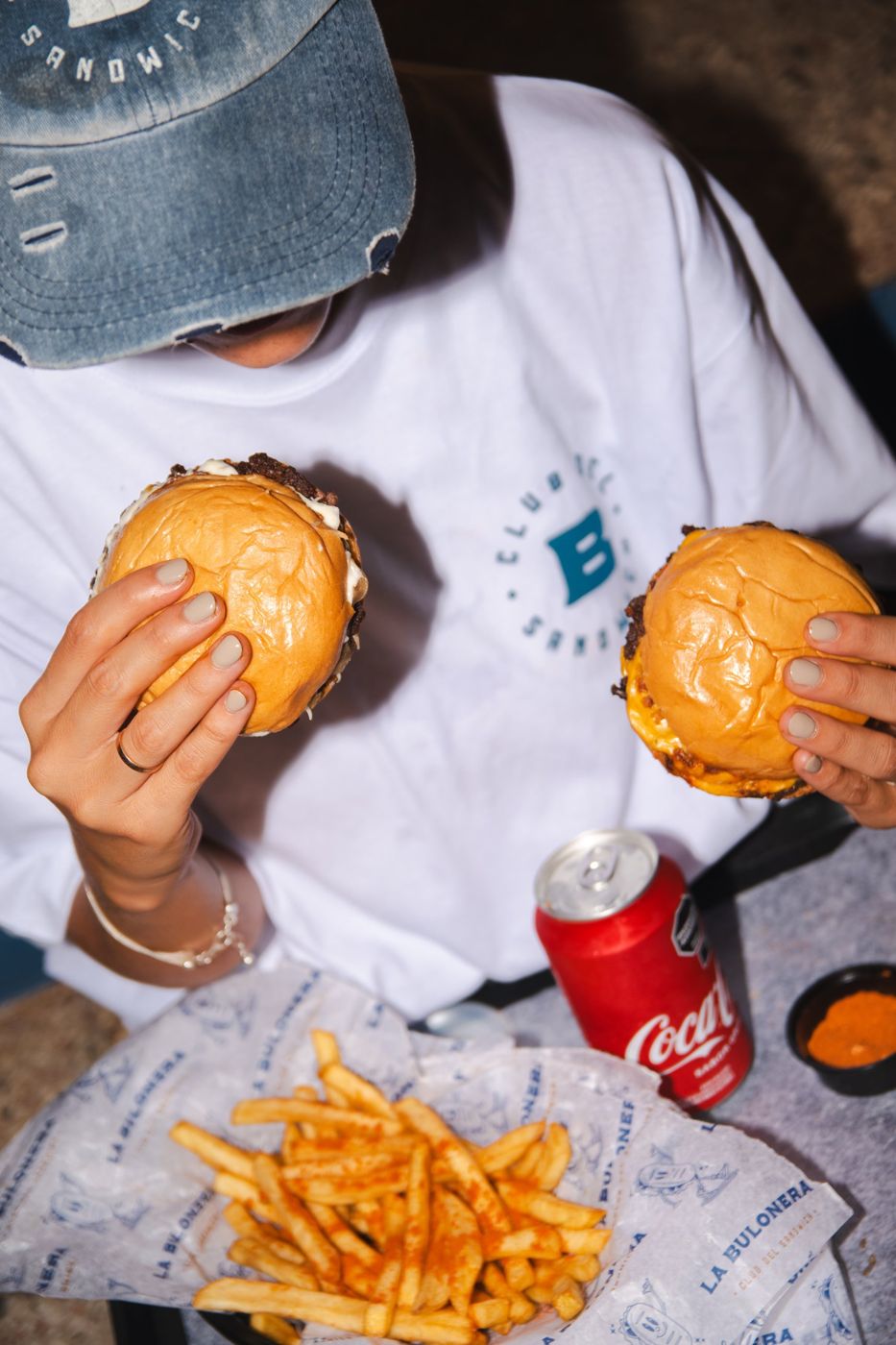 Una persona con remera de La Bulonera sostiene dos hamburguesas sobre una canasta de papas fritas